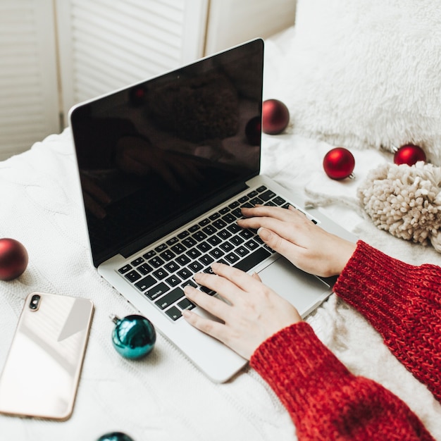 Mulher jovem com um suéter de malha vermelha digitando no laptop na cama branca com um cobertor branco decorado com bolas vermelhas e azuis de Natal