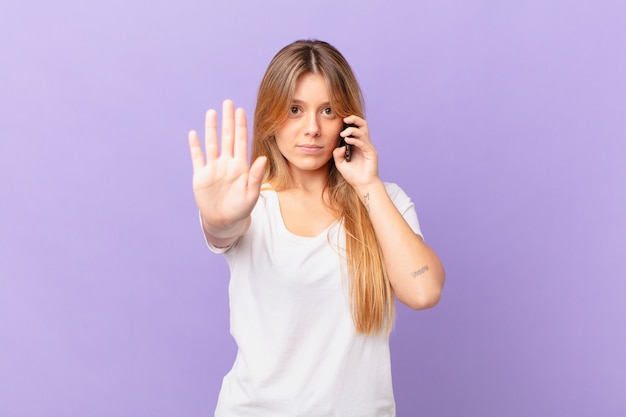 Mulher jovem com um telefone celular parecendo séria, mostrando a palma da mão aberta fazendo gesto de pare