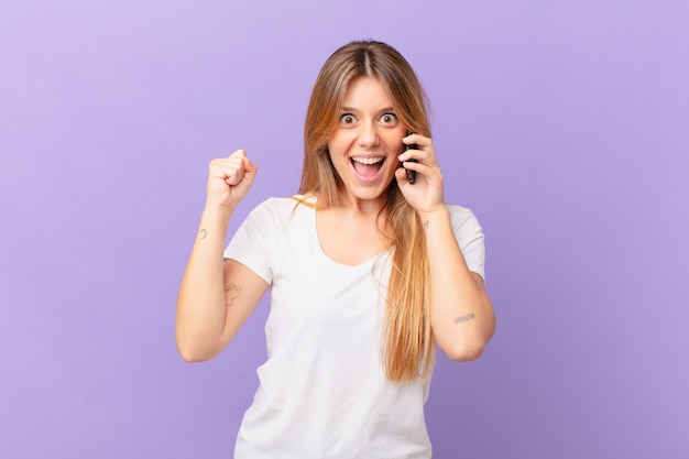 Mulher jovem com um telefone celular se sentindo chocada, rindo e comemorando o sucesso