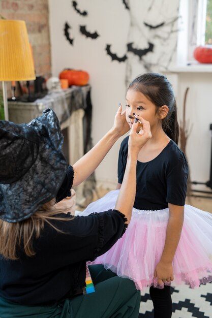 Mulher jovem com um traje preto de halloween, aplicando maquiagem no rosto de uma menina