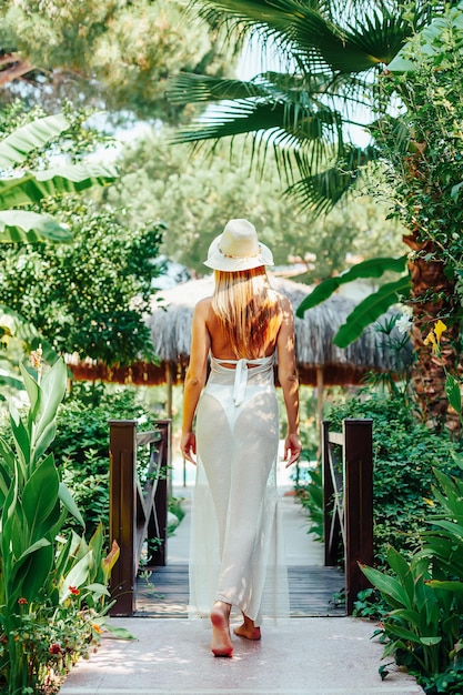 Mulher jovem com um vestido longo e um chapéu, caminhando pelo belo jardim tropical verdejante