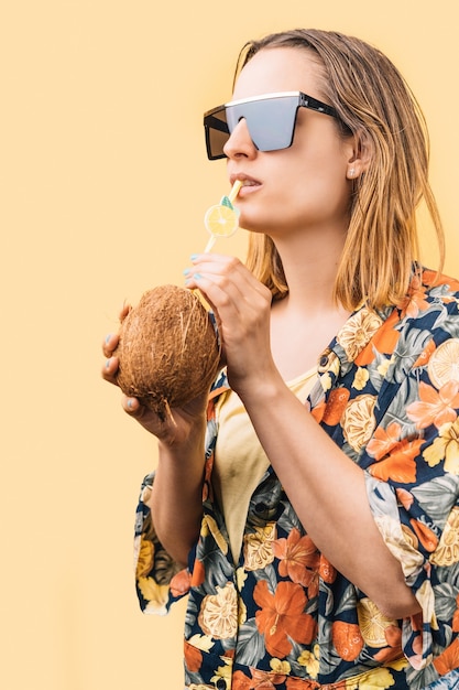 Mulher jovem com uma camisa florida e grandes óculos escuros bebendo um coquetel de coco com um canudo