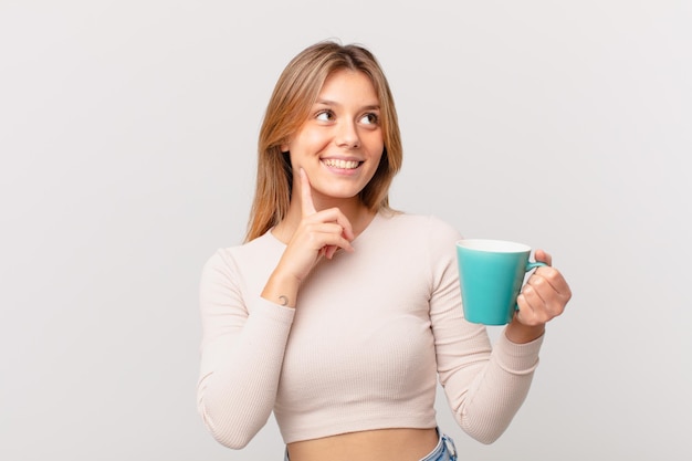 Mulher jovem com uma caneca de café sorrindo feliz e sonhando acordada ou duvidando