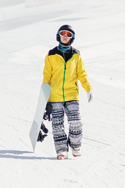 Mulher jovem com uma prancha de snowboard na mão caminhando em uma pista de esqui, ela está olhando para a câmera, copie o espaço