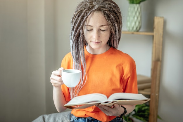 Mulher jovem com uma xícara de café ou chá e roupas brilhantes está sentada e lendo um livro em casa