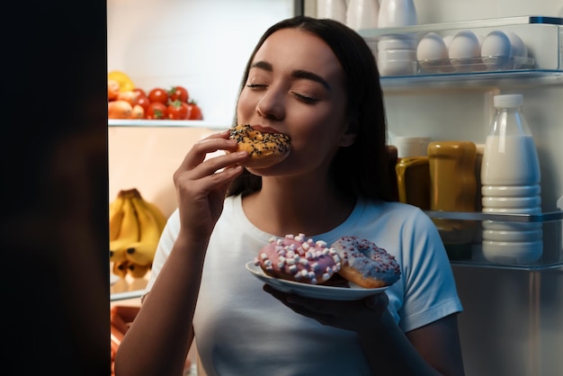 Mulher jovem comendo donut perto da geladeira na cozinha à noite Mau hábito
