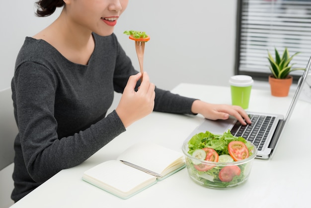 Mulher jovem comendo salada enquanto trabalhava na mesa. Conceito de perda de peso