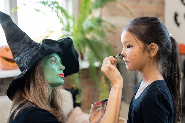 Mulher jovem concentrada com bigodes verdes pintando o rosto das crianças enquanto a prepara para a celebração do Halloween