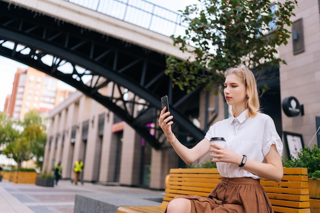 Mulher jovem confiante tomando selfie usando smartphone e segurando xícara com café para viagem em dia de verão, sentado no banco na rua da cidade