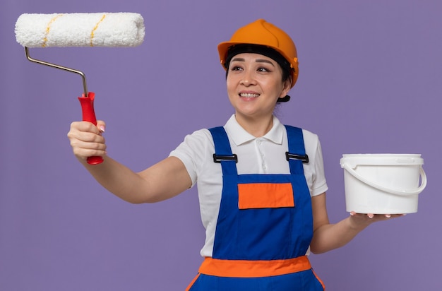 Mulher jovem construtora asiática sorridente com capacete laranja olhando para o rolo de pintura e segurando tinta a óleo