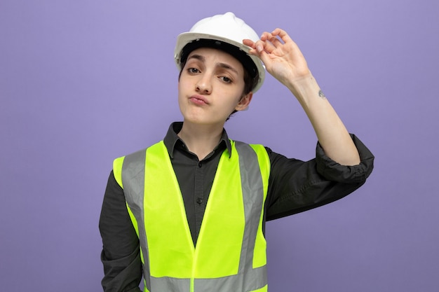 Mulher jovem construtora em colete de construção e capacete de segurança olhando com expressão séria e confiante tocando seu capacete