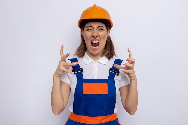 Mulher jovem construtora em uniforme de construção e capacete de segurança, gritando e gritando com os braços levantados, sendo frustrada e louca louca de pé no branco