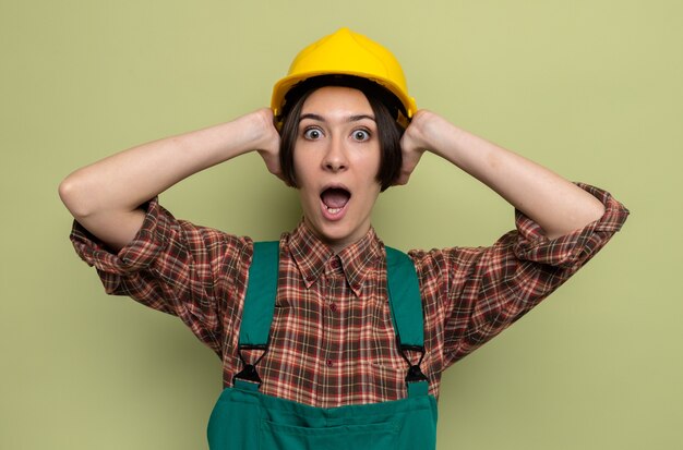 Mulher jovem construtora em uniforme de construção e capacete de segurança olhando para a frente preocupada e chocada, segurando as mãos na cabeça em pânico em pé sobre a parede verde