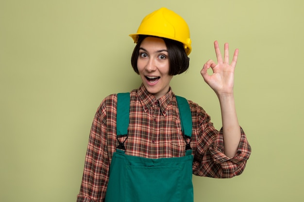 Mulher jovem construtora feliz em uniforme de construção e capacete de segurança sorrindo alegremente fazendo sinal de ok em pé no verde