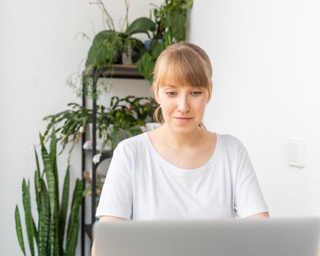 Mulher jovem conversando na varanda do laptop com plantas em vasos de fundo interior