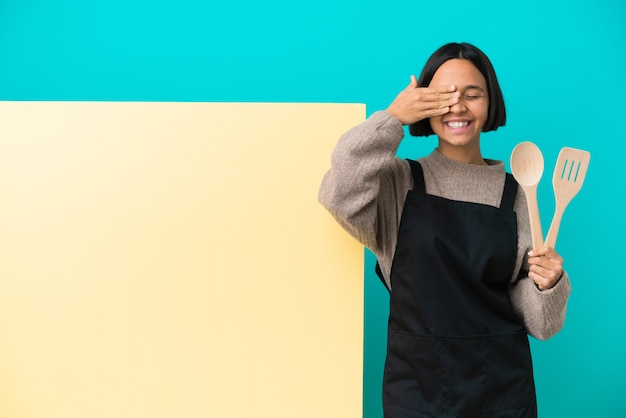 Mulher jovem cozinheira de raça mista com um grande cartaz isolado em um fundo azul cobrindo os olhos pelas mãos e sorrindo