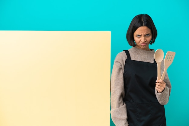 Mulher jovem cozinheira de raça mista com um grande cartaz isolado em um fundo azul com uma expressão infeliz
