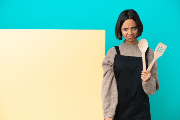 Mulher jovem cozinheira de raça mista com um grande cartaz isolado em um fundo azul com uma expressão triste