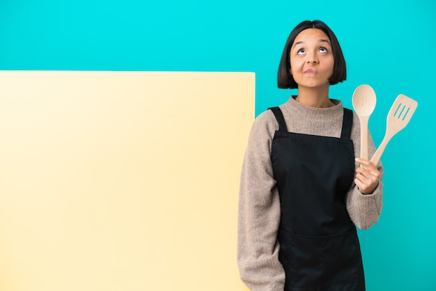 Mulher jovem cozinheira de raça mista com um grande cartaz isolado em um fundo azul e olhando para cima