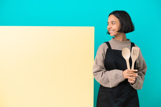 Mulher jovem cozinheira de raça mista com um grande cartaz isolado em um fundo azul, olhando de lado