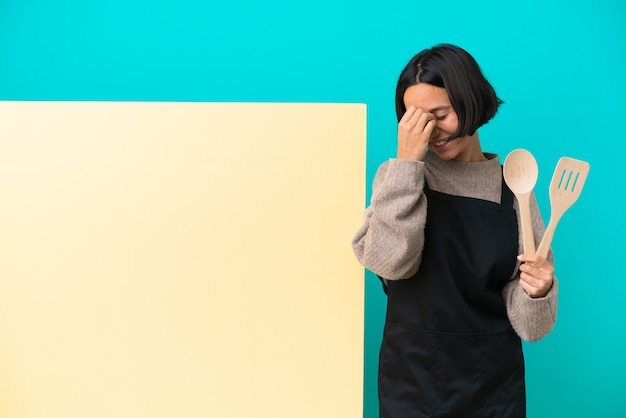 Foto mulher jovem cozinheira de raça mista com um grande cartaz isolado em um fundo azul rindo