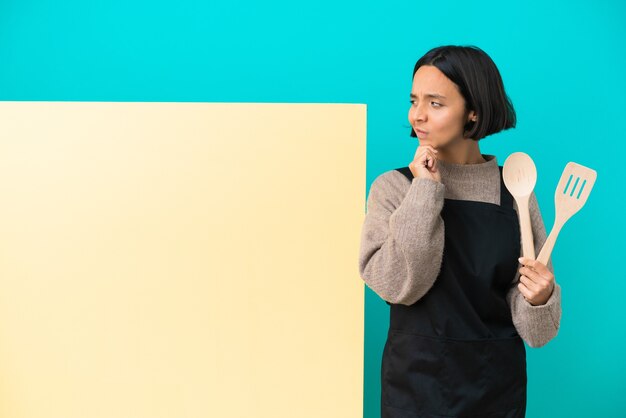 Mulher jovem cozinheira de raça mista com um grande cartaz isolado em um fundo azul, tendo dúvidas e pensando