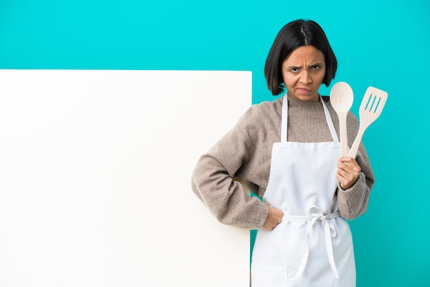 Mulher jovem cozinheira de raça mista com um grande cartaz isolado na parede azul com raiva