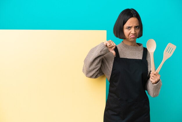 Foto mulher jovem cozinheira de raça mista com um grande cartaz isolado na parede azul, fazendo sinais de bom-mau. indeciso entre sim ou não