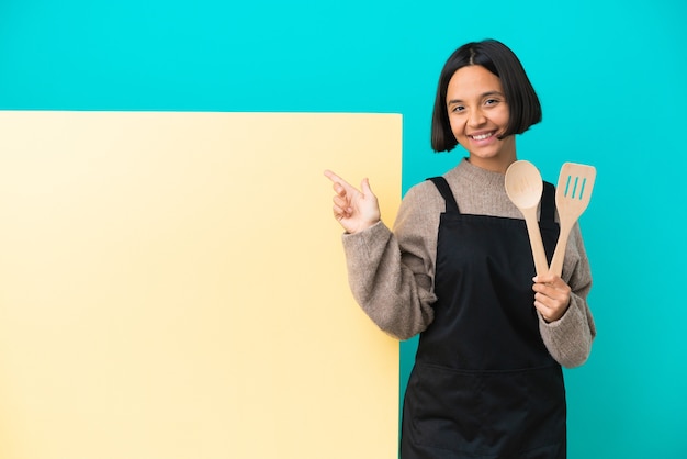 Mulher jovem cozinheira de raça mista com um grande cartaz isolado na parede azul feliz e apontando para cima