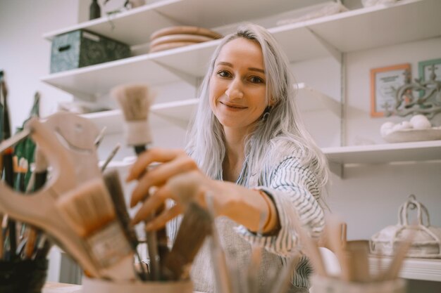 Mulher jovem criando produto de barro em uma oficina de estúdio de arte Menina sorridente usando avental fazendo utensílios de mesa na aula de cerâmica Artesã usando ferramenta manual trabalhando em objeto de cerâmica em uma oficina de cerâmica