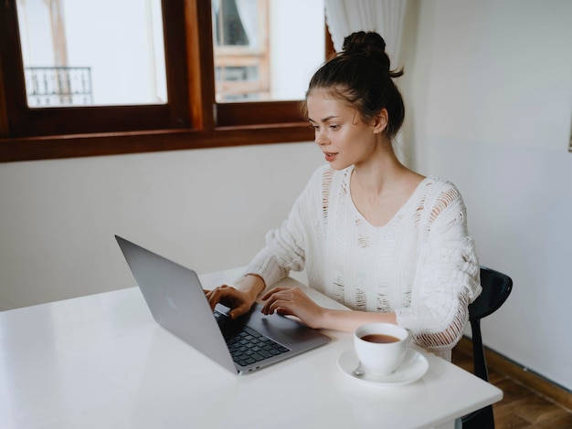 Mulher jovem criativa trabalhando em uma mesa em casa em um interior aconchegante com uma xícara de bebida home office freelancer com um sorriso comunicação e educação online