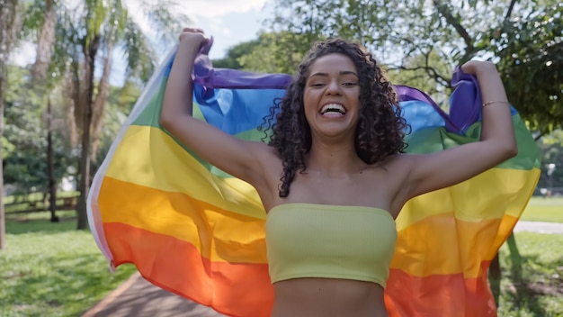 Mulher jovem de cabelo encaracolado cobrindo com a bandeira do orgulho lgbt. Mantendo o punho erguido, cobrindo a bandeira LGBT +