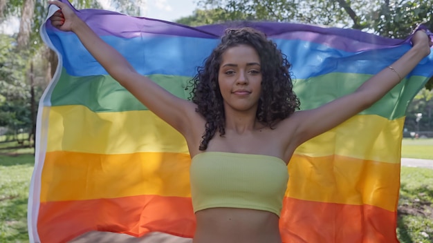 Mulher jovem de cabelo encaracolado cobrindo com a bandeira do orgulho lgbt. Mantendo o punho erguido, cobrindo a bandeira LGBT +