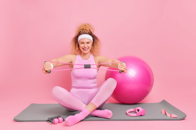 Mulher jovem de cabelo encaracolado feliz sentada com as pernas cruzadas no tapete de fitness treina os músculos