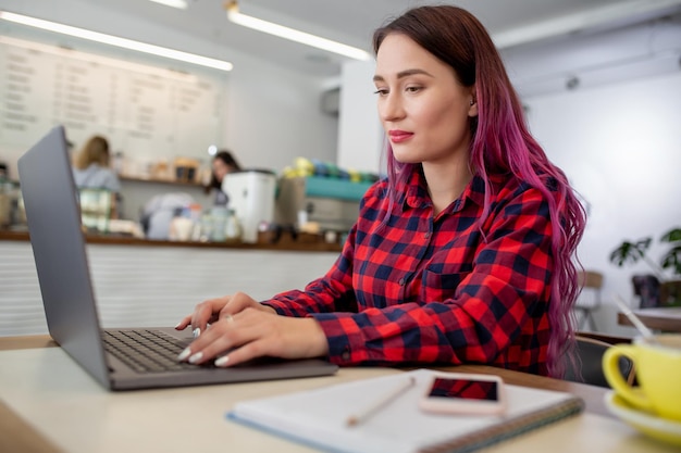 Mulher jovem de cabelo rosa com laptop sentada em um café, estudante inteligente trabalhando no netbook após suas palestras na universidade