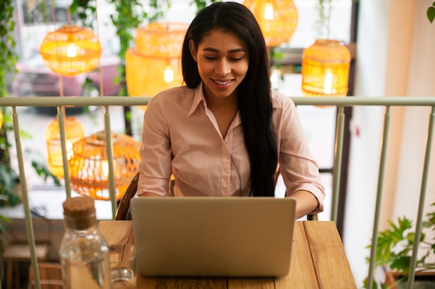 Mulher jovem de cabelos compridos parecendo entusiasmada enquanto está sentada no café com lindas lâmpadas nas costas e trabalhando no laptop