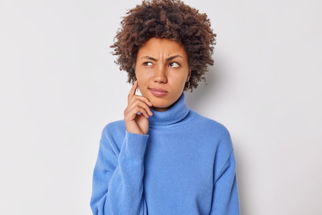 Mulher jovem de cabelos encaracolados séria considera que algo pondera sobre planos futuros mantém o dedo na bochecha usa jumper azul casual isolado sobre o fundo branco do estúdio. deixe-me pensar o que decidir