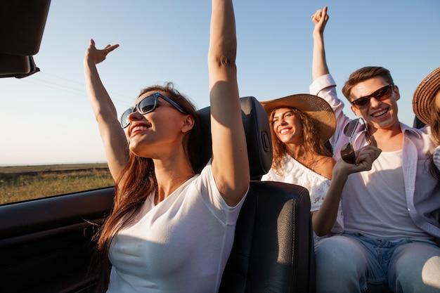 Mulher jovem de cabelos escuros, vestida com uma camiseta branca, está sentada em um cabriolet preto com amigos em um dia de verão. .