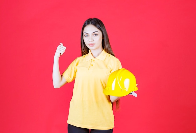 Mulher jovem de camisa amarela segurando um capacete amarelo e apontando para alguém atrás