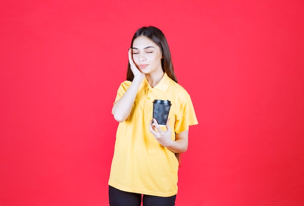 Mulher jovem de camisa amarela segurando uma xícara de café descartável preta e parece cansada e com sono