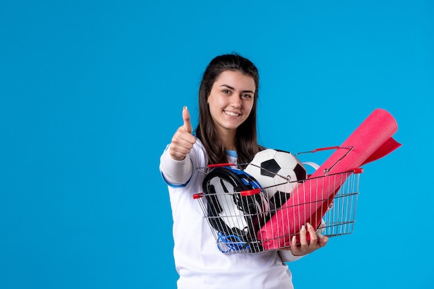 Mulher jovem de frente com uma cesta cheia de coisas esportivas na parede azul