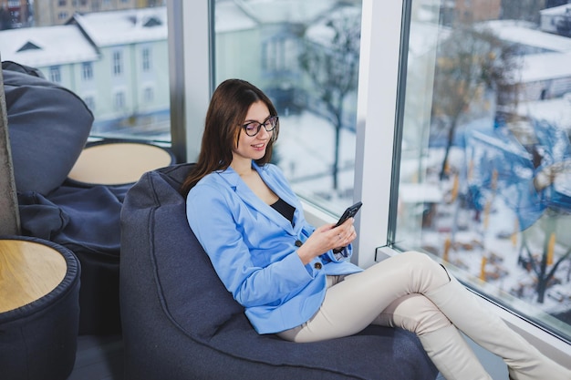 Mulher jovem de negócios em óculos com longos cabelos escuros em roupas casuais sorrindo e olhando para o telefone navegando no smartphone durante um dia de folga no espaço de trabalho