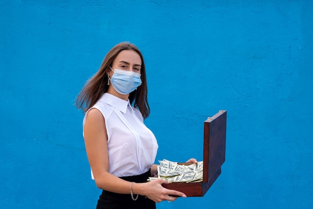 Mulher jovem de negócios usar máscara respiratória segurando notas de dólar isoladas na parede azul. conceito de quarentena em casa, prevenção covid-19