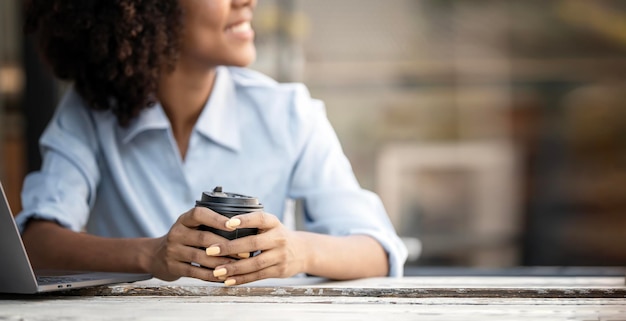 Mulher jovem de pele negra segurando a xícara de café sentada no espaço da cópia da mesa