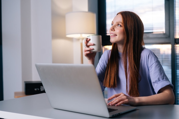 Mulher jovem de pensamento um sonho usando o computador portátil e bebendo café no local de trabalho em casa.