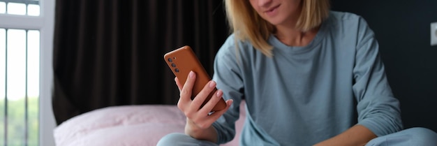 Mulher jovem de pijama sentada na cama com o celular na mão