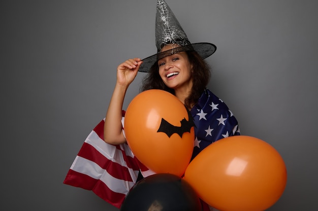 Mulher jovem de raça mista alegre com chapéu de mago, embrulhada na bandeira americana, segura bolas coloridas de ar laranja, sorri, sorriso cheio de dentes, olhando para a câmera. Conceito de Halloween em fundo cinza com espaço de cópia