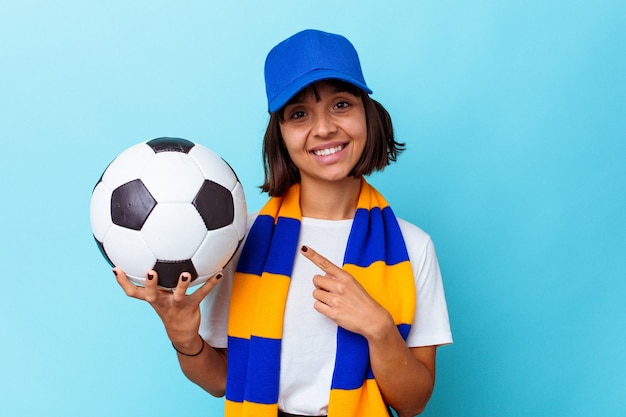 Mulher jovem de raça mista assistindo futebol isolado em fundo azul, sorrindo e apontando de lado, mostrando algo no espaço em branco.