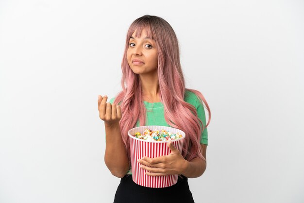 Mulher jovem de raça mista com cabelo rosa comendo pipoca isolada no fundo branco fazendo gesto de dinheiro