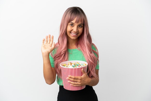 Mulher jovem de raça mista com cabelo rosa comendo pipoca isolada no fundo branco saudando com a mão com expressão feliz